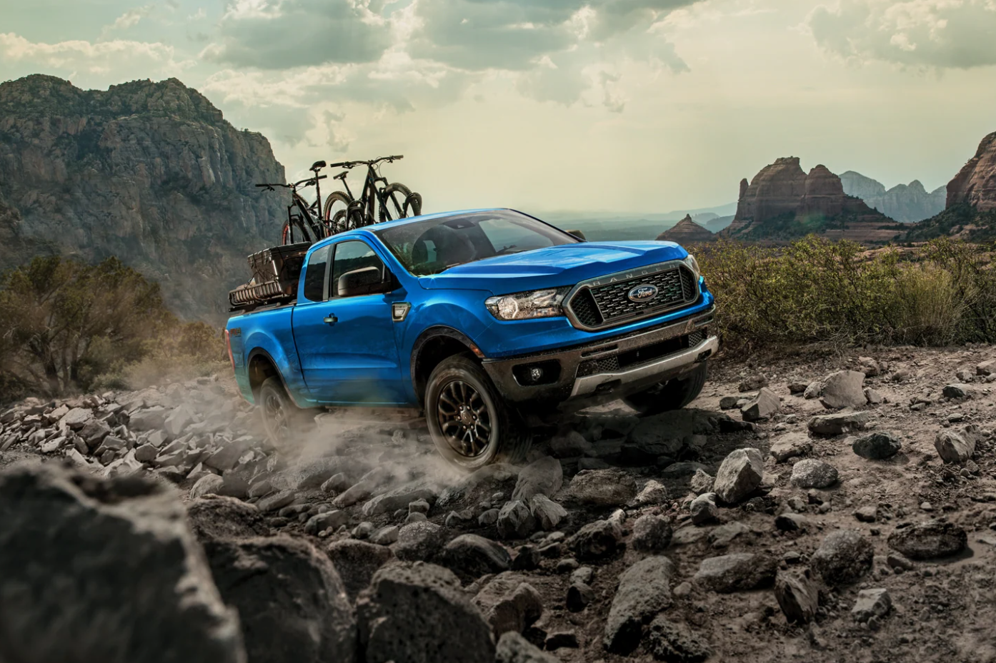 A bright blue 2022 Ford Ranger drives across a rocky terrain, kicking up dust and hauling mountain bikes in the truck bed in a mountainous background