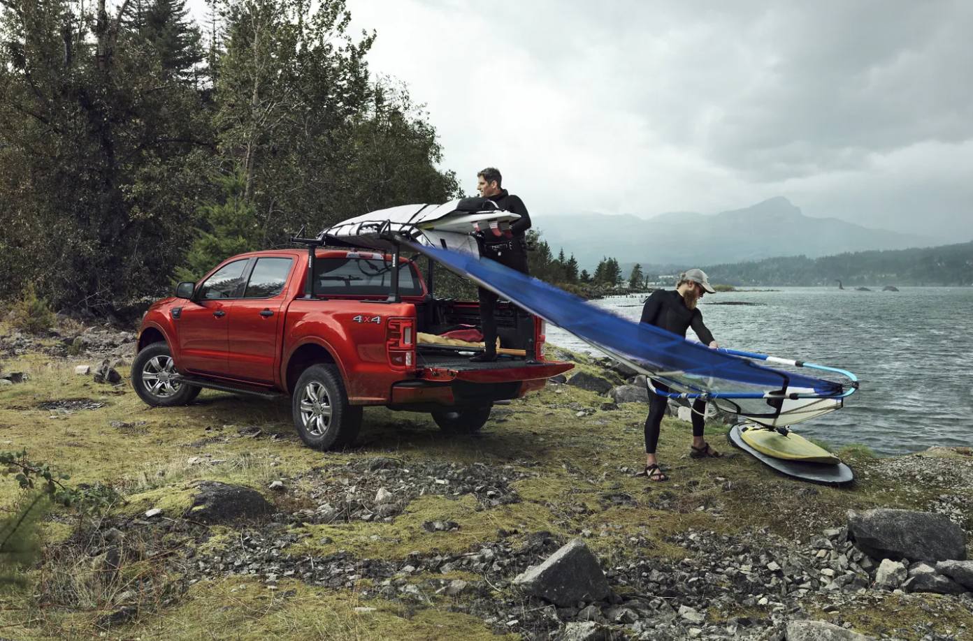 A red 2022 Ford Ranger sits parked next to a lake, facing away from the viewer as two people unload gear from the bed of the truck