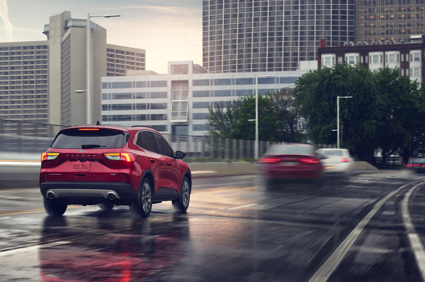 A view of the back of a red 2022 Ford Escape as it drives down a wet street after a rainstorm towards a city skyline