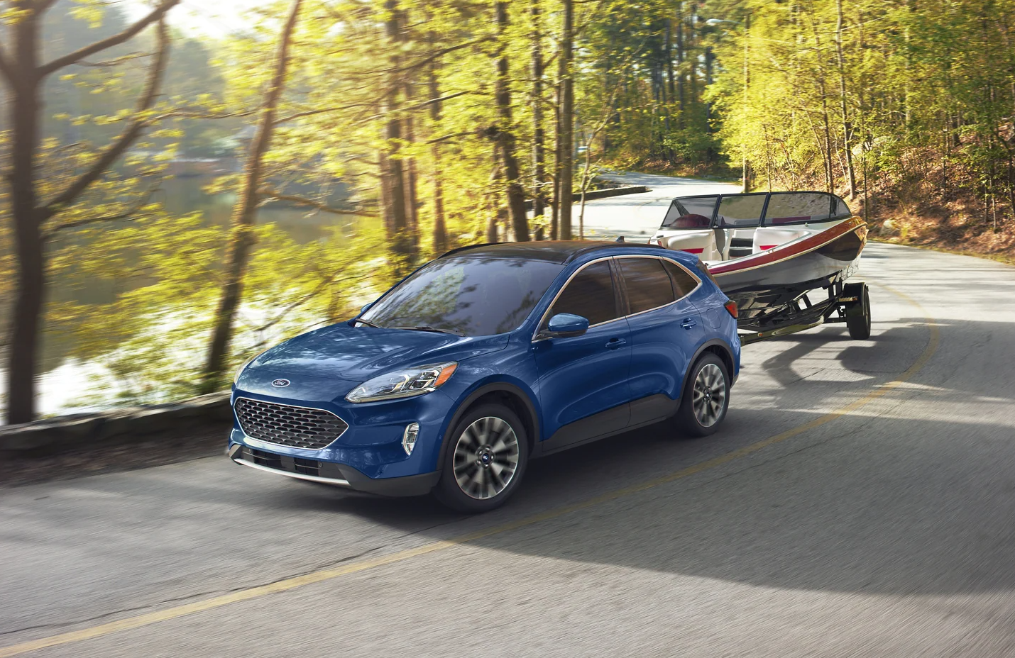 alt="A medium blue 2022 Ford Escape hauls a speedboat down a sun dappled road edged with trees and a lake on the left"