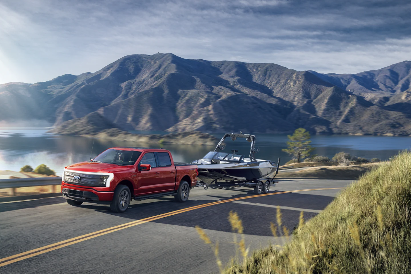 A red 2023 Ford F-150 Lightning cruises down a highway next to a mountain lake hauling a boat