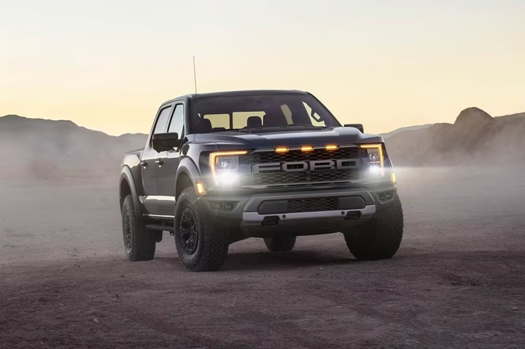 A black 2023 Ford Ranger Raptor sits parked in a dusty field with its headlights on