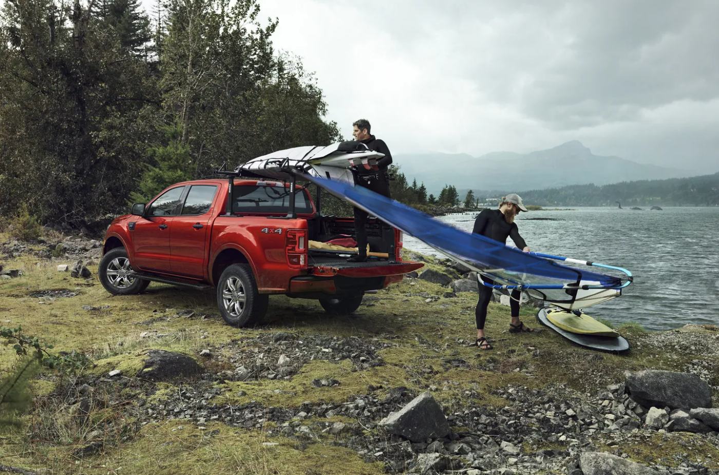 Two men unload windsurfing gear from the back of a red 2023 Ford Ranger parked next to a lake